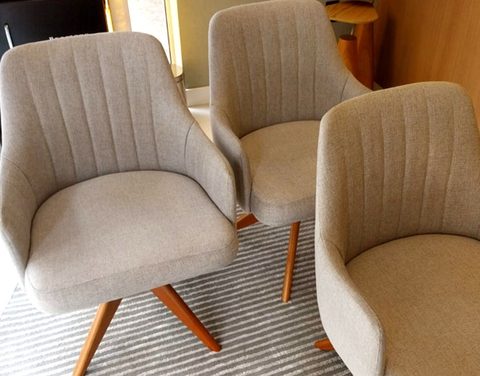 Três poltronas giratórias de tecido bege com pés de madeira, sobre tapete listrado, em uma sala junto à janela