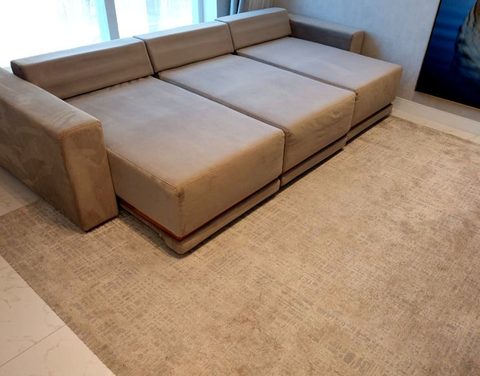 Sofá retrátil de suede bege, com três assentos e chaise, com os assentos avançados, sobre tapete claro em sala de apartamento